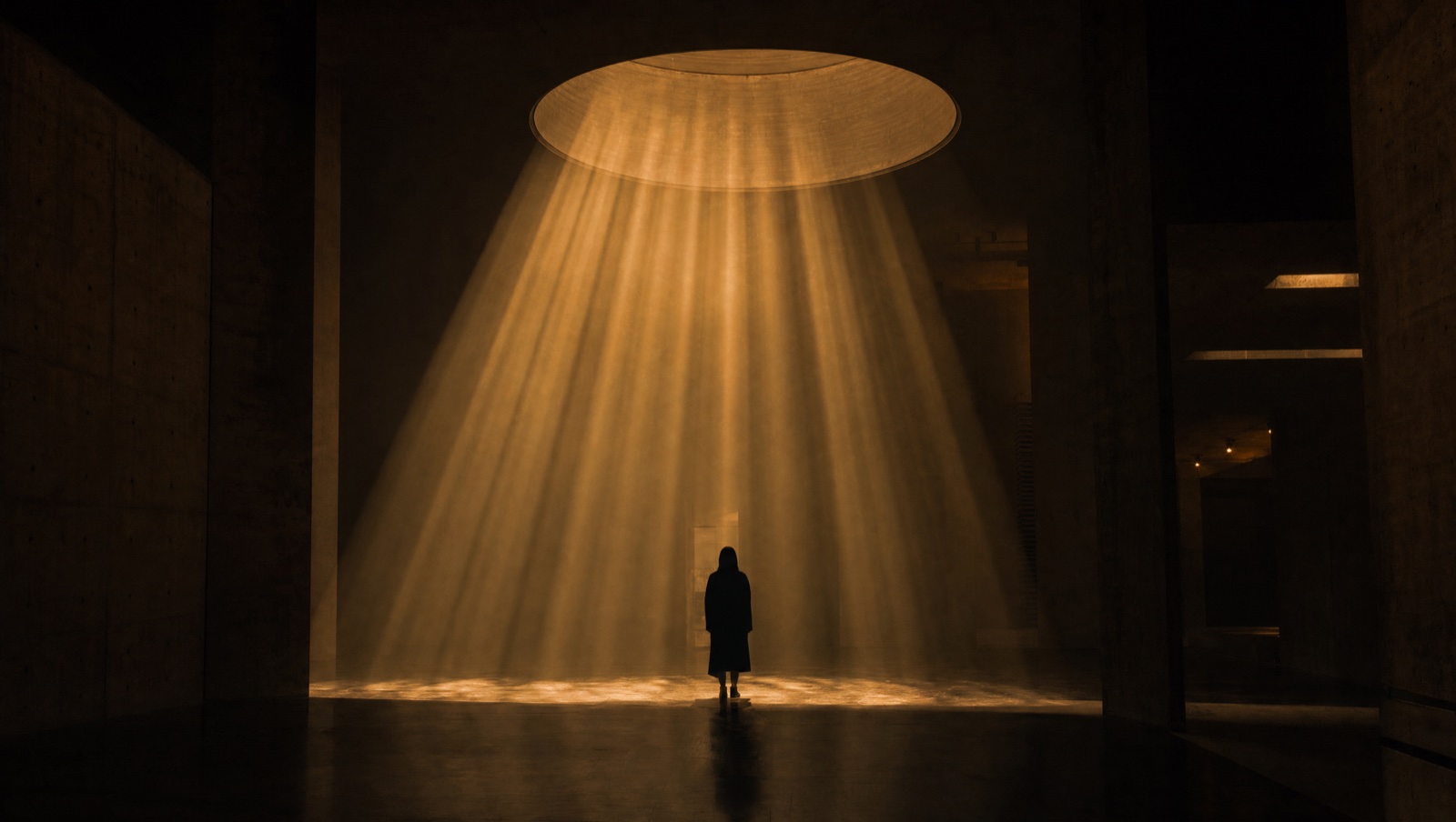A lone visitor stands inside a cone of amber volumetric light in a dark concrete hall.