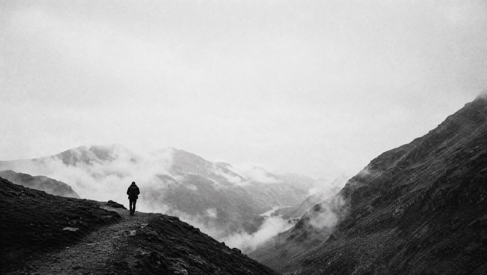 Black and white foggy mountain valley with a single walker on a ridge path.